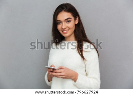 Charming smiling girl holding mobile phone and looking at camera, isolated over gray background