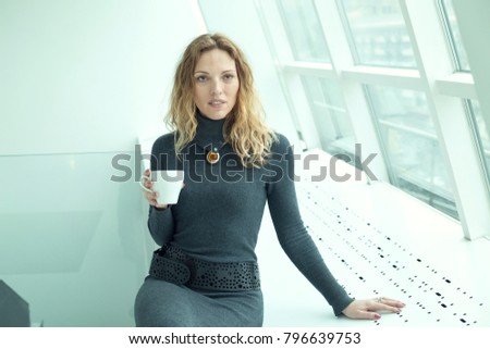 Woman is holding a cup of coffee while standing in the office near the window.