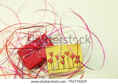 A red little gift with a yellow post-it with hand painted hearts lie on a white background.