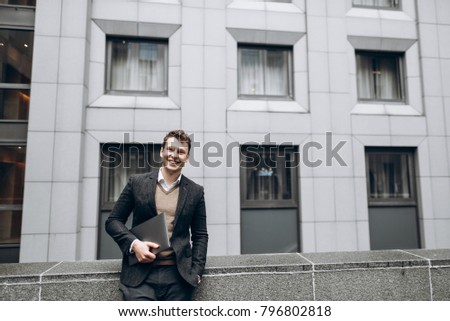 Image of a smiling handsome businessman with a laptop in his hands who stands outside in the background of a business center