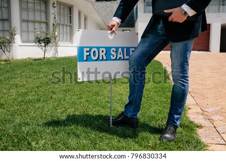 Realtor with a for sale sign board outside a house. Male Real Estate broker with for sale notice in lawn of a new house.