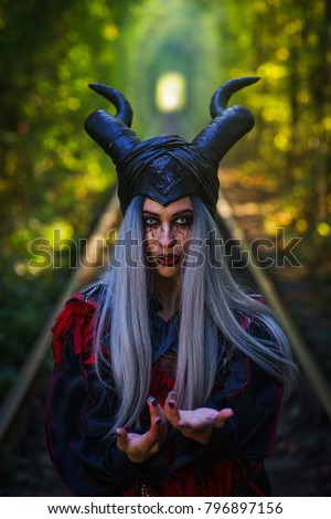 terrible woman with horns and amazing makeup in the tunnel of forest with rails close up