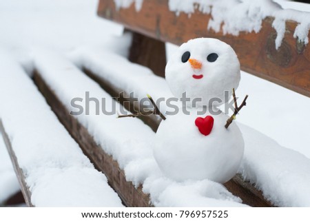 Happy little snowman with a red heart on a snowy bench