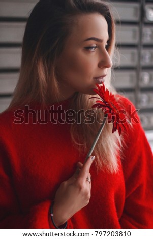 woman with a red flower - gerbera on the background of postal cells. she is dressed in a red top or sweater. hair dyed ombre, blonde and brunette. girl with makeup.