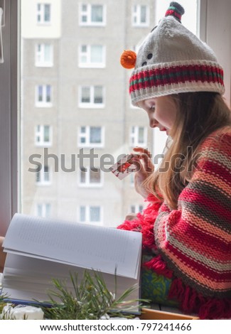 A winter photo of a little girl in a knitted sweater sitting on a windowsill and reading a book