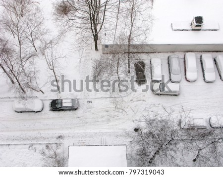 Winter, all in snow. Top view on inside road of residential courtyard road in city. Cars in parking are covered snow. Bare trees.