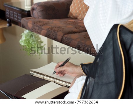 Saudi Arabian Man Hand Writing on A Notebook in a Luxury Home Environment, wearing Saudi Thob, Ghutra and Black Bisht