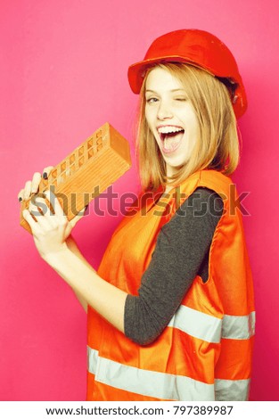 Excited pretty girl or young cute beautiful woman or construction worker in hard hat and reflective jacket winks with red brick on pink background