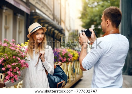 Tourist Couple. Man With Camera Taking Photos Of Woman In Street. Smiling Young Couple In Love In Stylish Clothes Traveling And Making Photos Outdoors. People Travel On Weekend. High Resolution.