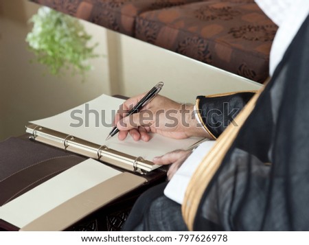 Saudi Arabian Man Hand Writing on A Notebook in a Luxury Home Environment, wearing Saudi Thob, Ghutra and Black Bisht