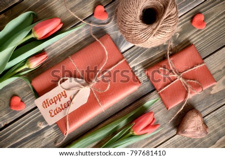 Easter arrangement: red tulips, wrapped gifts, cord and decorative hearts on wooden tabke, flat lay. Words on the tag: "Happy Easter!"
