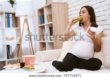 Pregnant girl eating a hamburger and chocolate sitting on the couch. She's at home. She is hungry.
