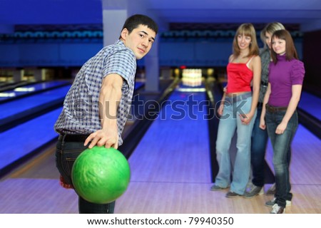 Young man prepares throw ball on path for bowling and three girls look on him, focus on man