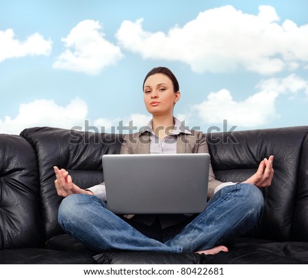 Portrait of young business woman sitting with a laptop on comfortable black couch and meditating on cyan sky background