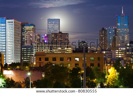 An eerie glowing moon rises  behind a tall skyscraper in the Denver Colorado skyline.