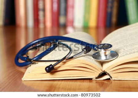 A stethoscope is lying with a book on the desk against books Royalty-Free Stock Photo #80973502