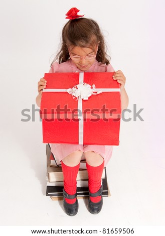 A little girl sitting on a pile of books opening the lid of a big present