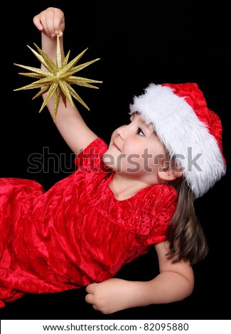 little girl in Santa hat laying down and looking up at the golden Christmas ornament in her hand. isolated on black