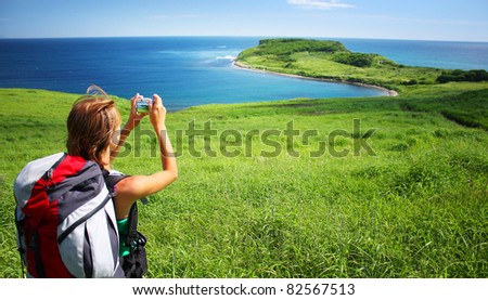 Young woman with backpack standing on a hill with green grass and making snapshot of a beautiful scene