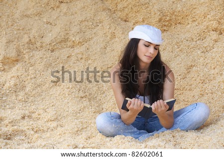 Portrait of young beautiful woman wearing white cap, t-shirt and jeans sitting in sawdust and reading book against yellow background.