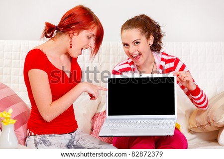 Two cheerful girlfriends pointing on laptops blank screen  at living room