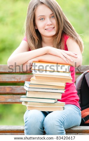 beautiful and happy young student girl sitting on bench, her hands on pile of books, looking into the camera and smiling. Summer or spring green park in background