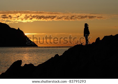 A woman standing on a rocky coastline, silhouetted by a sunset, in Norris Point, Newfoundland, Canada.