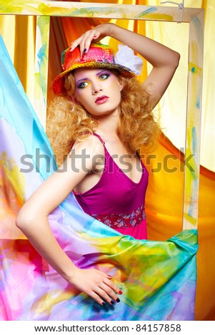 portrait of beautiful blonde woman artist in colorful hat standing behind the easel with painted cloth on it