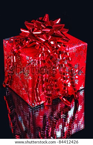 A Christmas gift wrapped in metallic Christmas paper and decorated with a red foil bow and ribbon on a dark background.
