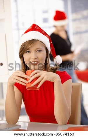 Female office worker sitting at office desk, wearing xmas hat, drinking coffee.?