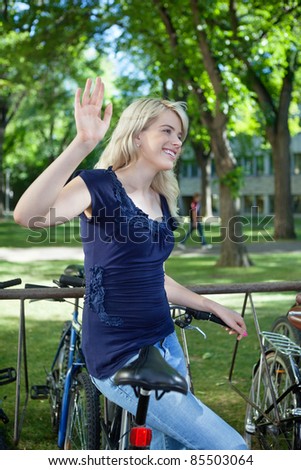 College student waving hand at someone while standing near by her bike