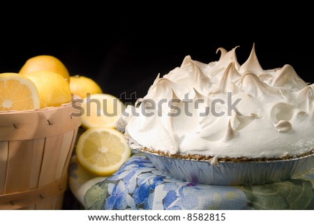 Still life of lemon meringue pie next to basket of lemons on blue print tablecloth