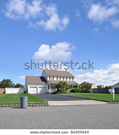 Recycle Can in Front of Residential Suburban High Ranch Home with Two Car Garage Blacktop Driveway Mailbox on Front Lawn Sunny Blue Sky with Clouds