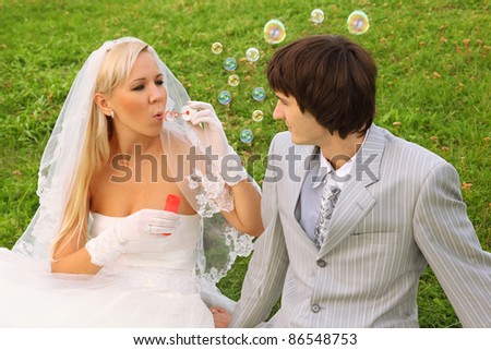 Beautiful young bride wearing white dress sitting on green grass with groom and blowing bubbles; focus on man