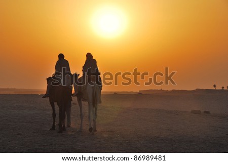 two camels riding towards the sun in the desert Royalty-Free Stock Photo #86989481
