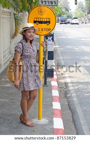 Young woman standing at a bus stop on street of city. Asian woman waiting for the bus. Woman in summer dress standing at the bus stop. Public transport stop in Thailand. Signs bus stops on the street.