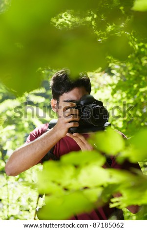 young hispanic man trekking among trees and taking pictures with dslr camera. Vertical shape, front view, copy space