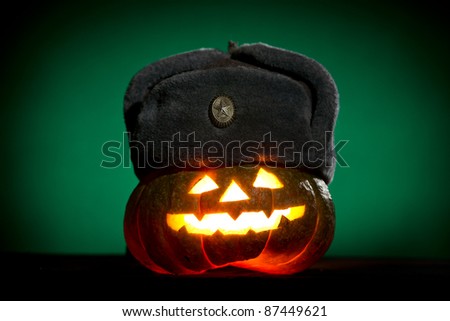 Orange scarry pumpkin with burning eyes, nose and mouth with winter military hat on green background in the dark