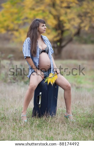 Pregnant woman in the autumn park. Expectant mother in unbuttoned checkered shirt and shorts sits on valise