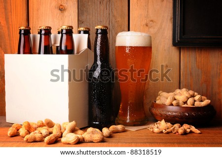  A glass of beer next to a six pack in a rustic tavern setting. Shelled peanuts in a bowl and strewn on the wood table surface. A blank picture frame hangs on the wall ready for your type or image.