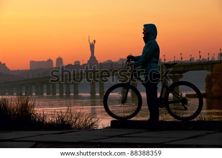 Young woman with her bicycle standing on river bank against sunset sky