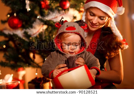 Young mother helping interested baby open present box at Christmas tree