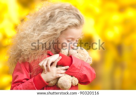 Beautiful and happy little girl with teddy bear on her hands. Kissing her toy with her eyes closed. Yellow autumn trees in background.