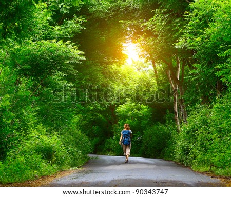 Woman with backpack walking on an asphalt road in a park with green trees