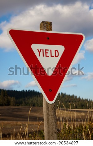 A new red yield sign is bolted to an old mossy post on a country road with grassy fields and tall trees in the distance.