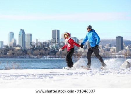 Winter fun couple on snowshoes running with montreal cityscape skyline and river st. Lawrence in background. Healthy lifestyle photo from Montreal, Quebec, Canada.
