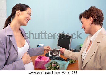 Photo of a pregnant women at work in her office showing her baby ultrasound scan print to a colleague, flowers and gift on the desk as she goes on maternity leave.