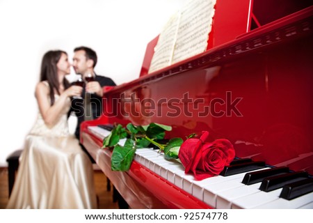 Red rose on the red grand piano keyboard in focus and couple sitting on the chair with glasses of wine at background