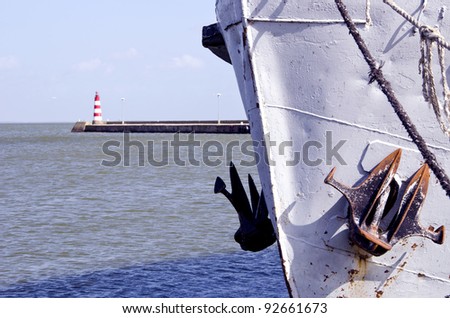 Ship fore with raised anchor closeup and red lighthouse in distance.