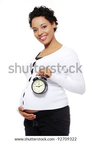 Site view of a beautiful young woman holding an alarm clock in front of her tummy, isolated on a white background.